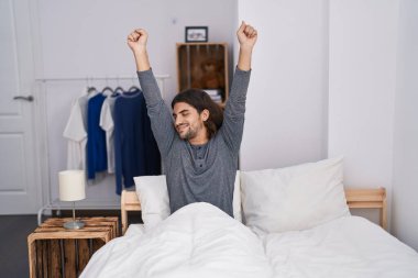Young hispanic man waking up stretching arms at bedroom