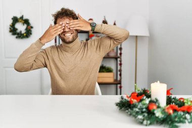 Masaya sakallı genç yakışıklı bir adam oturuyor Noel süslemesi ile gözleri kapayarak neşeli ve komik gülümsüyor. kör kavram. 