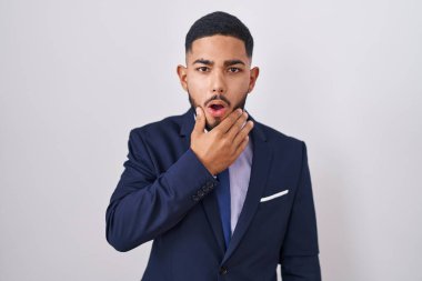 Young hispanic man wearing business suit and tie looking fascinated with disbelief, surprise and amazed expression with hands on chin 