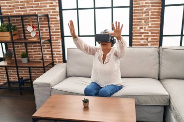 Evde sanal gerçeklik gözlüğü kullanan orta yaşlı bir kadın video oyunu oynuyor.