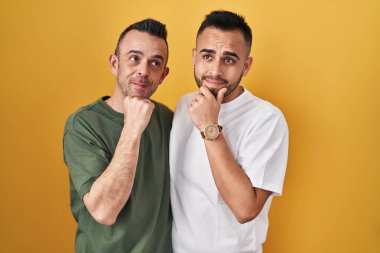 Homosexual couple standing over yellow background with hand on chin thinking about question, pensive expression. smiling and thoughtful face. doubt concept. 