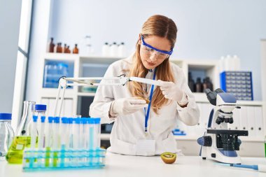 Bilim adamı üniforması giyen genç sarışın kadın laboratuarda büyüteç kullanarak kiviyi inceliyor.