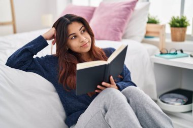 Young hispanic woman reading book sitting on floor at bedroom