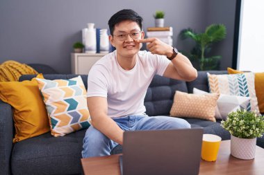 Young asian man using laptop at home sitting on the sofa pointing with hand finger to face and nose, smiling cheerful. beauty concept 