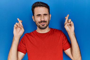Young hispanic man with beard wearing red t shirt over blue background gesturing finger crossed smiling with hope and eyes closed. luck and superstitious concept. 
