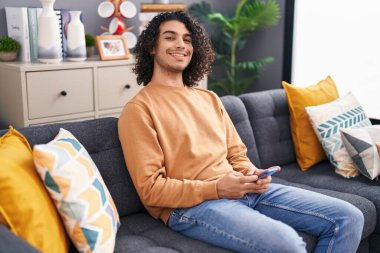 Evdeki kanepede oturan akıllı telefon kullanan genç latin adam.