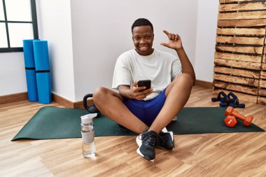 Young african man sitting on training mat at the gym using smartphone smiling and confident gesturing with hand doing small size sign with fingers looking and the camera. measure concept. 