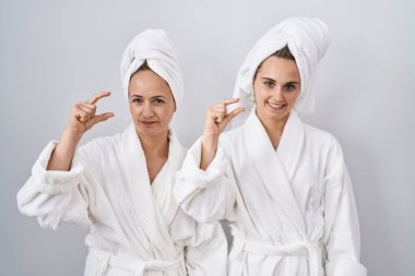 Middle age woman and daughter wearing white bathrobe and towel smiling and confident gesturing with hand doing small size sign with fingers looking and the camera. measure concept. 