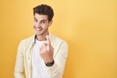 İspanyol asıllı genç adam sarı arka planda duruyor. Buraya el kol hareketi yaparak mutlu bir şekilde karşılamaya ve gülümsemeye davet ediyor. 
