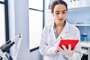 Laboratuvarda dokunmatik ped kullanan genç İspanyol kadın bilim adamı üniforması giyiyor.