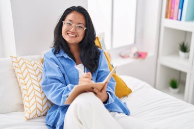 Young chinese woman writing on notebook sitting on bed at bedroom