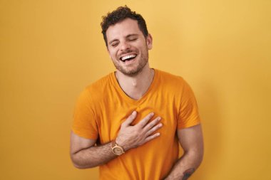 Sarı arka planda dikilen Latin asıllı genç adam gülümseyerek kahkahalarla gülüyor.. 