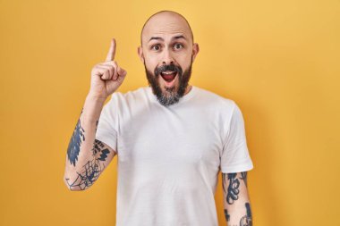 Sarı arka planda dikilen dövmeleri olan İspanyol genç adam başarılı bir fikri işaret ediyor. Heyecanlı ve mutlu. Bir numara.. 