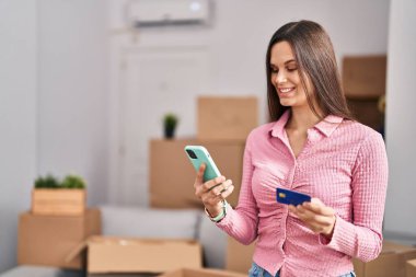 Young beautiful hispanic woman using smartphone and credit card at new home