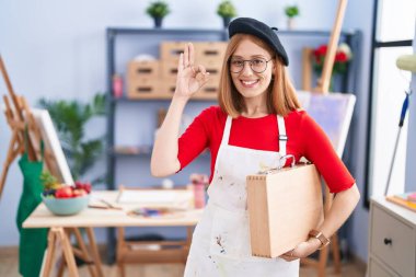 Young redhead woman at art studio holding art case smiling positive doing ok sign with hand and fingers. successful expression. 