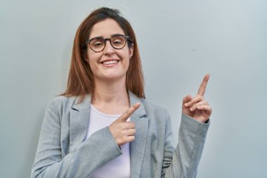 Gri arka planda duran esmer kadın gülümsüyor ve kameraya bakıyor iki eliyle ve parmaklarıyla kenarı işaret ediyor.. 