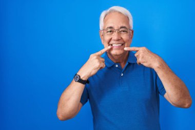 Mavi arka planda gri saçlı, neşeli gülümseyen ve parmaklarını ve ağzını gösteren orta yaşlı bir adam. diş sağlığı kavramı. 
