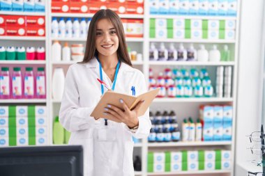 Young beautiful hispanic woman pharmacist smiling confident writing on notebook at pharmacy