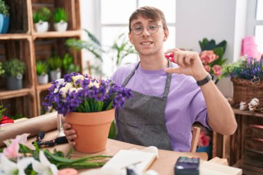 Caucasian blond man working at florist shop smiling and confident gesturing with hand doing small size sign with fingers looking and the camera. measure concept. 