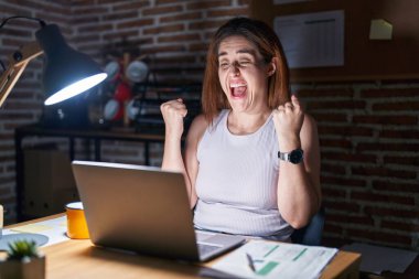 Geceleri ofiste çalışan esmer kadın kollarını kaldırıp, gözlerini açarak başarının şaşkınlığını ve şaşkınlığını yaşıyor. Kazanan konsept. 