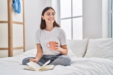 Young hispanic woman drinking cup of coffee reading book at bedroom