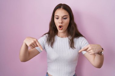Young hispanic girl standing over pink background pointing down with fingers showing advertisement, surprised face and open mouth 