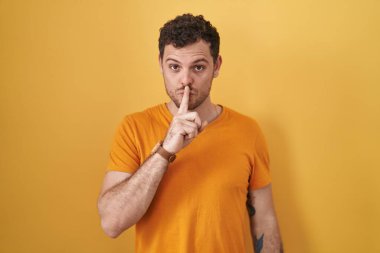 Sarı arka planda dikilen genç İspanyol adam parmakları dudaklarında sessiz olmak istiyor. sessizlik ve gizli konsept. 