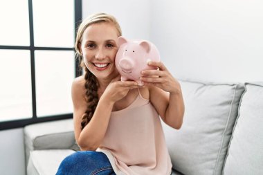 Young caucasian woman holding piggy bank sitting on sofa at home