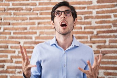 Young hispanic man standing over brick wall background crazy and mad shouting and yelling with aggressive expression and arms raised. frustration concept. 