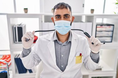 Young hispanic man doctor wearing medical mask holding stethoscope at clinic
