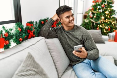 Young hispanic man using smartphone sitting on sofa by christmas tree at home