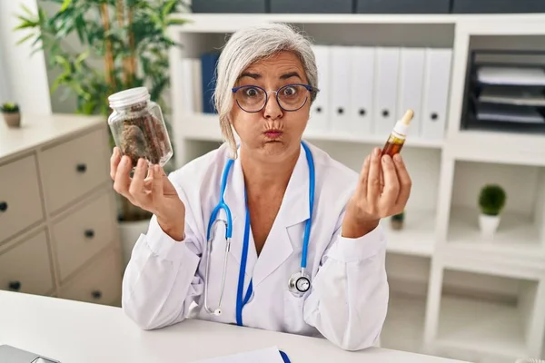 Middle age woman doctor holding cbd oil puffing cheeks with funny face. mouth inflated with air, catching air.