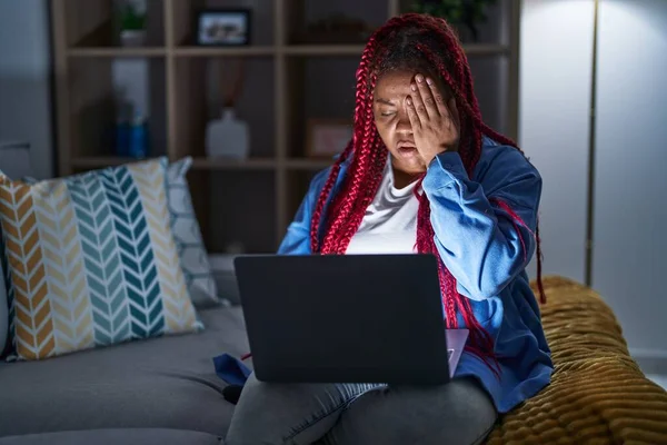 African american woman with braided hair using computer laptop at night yawning tired covering half face, eye and mouth with hand. face hurts in pain.