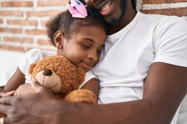Father and daughter hugging each other sitting on bed holding teddy bear at bedroom