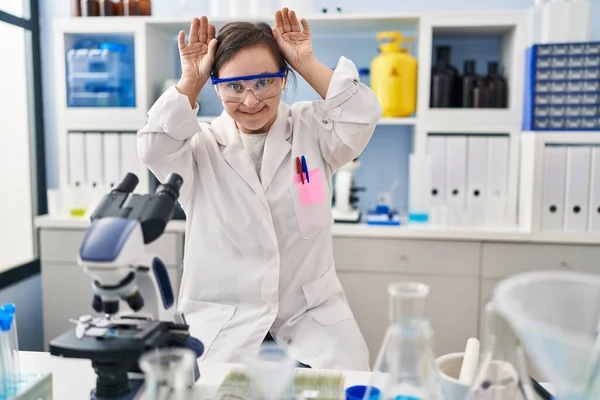 Hispanic girl with down syndrome working at scientist laboratory doing bunny ears gesture with hands palms looking cynical and skeptical. easter rabbit concept.