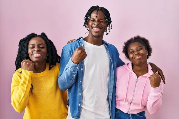 Group of three young black people standing together over pink background very happy and excited doing winner gesture with arms raised, smiling and screaming for success. celebration concept.