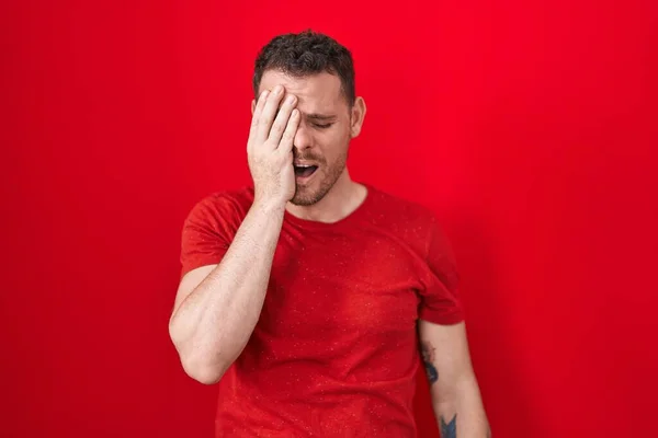 Young hispanic man standing over red background yawning tired covering half face, eye and mouth with hand. face hurts in pain.