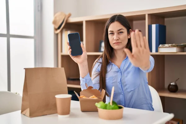 年轻的黑发女子在家里吃东西时 带着严肃而自信的表情和防守姿态 用张开的手展示智能手机屏幕 做停止手势 — 图库照片