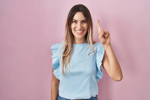Young hispanic woman standing over pink background showing and pointing up with finger number one while smiling confident and happy.