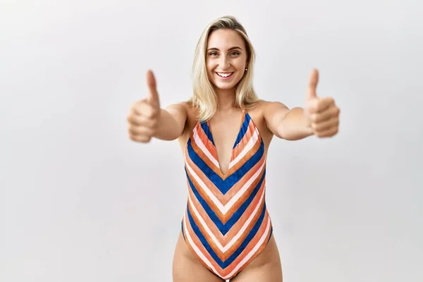 Young blonde woman wearing swimsuit over isolated background approving doing positive gesture with hand, thumbs up smiling and happy for success. winner gesture.