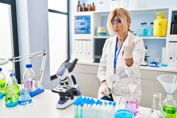 Middle age blonde woman working at scientist laboratory showing middle finger, impolite and rude fuck off expression