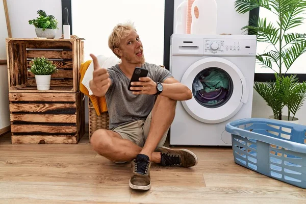 Young Blond Man Doing Laundry Using Smartphone Approving Doing Positive — Stok fotoğraf