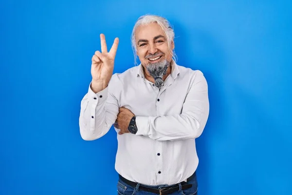 Middle age man with grey hair standing over blue background smiling with happy face winking at the camera doing victory sign with fingers. number two.