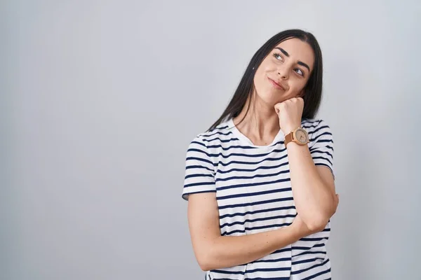 若いブルネットの女性は質問について考える顎の手で縞模様のTシャツを着て 集中的な表現 思慮深い顔で笑顔 疑わしい概念 — ストック写真