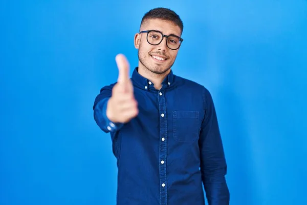 Young hispanic man wearing glasses over blue background smiling friendly offering handshake as greeting and welcoming. successful business.