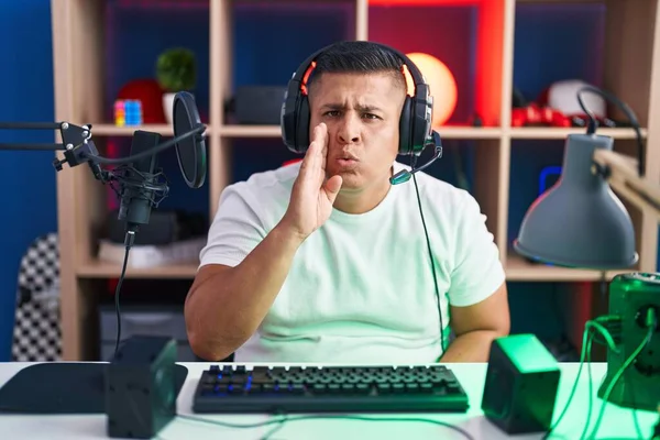 若いヒスパニック系の男がビデオゲームをしている手に口から秘密の噂を伝え 悪意のある話の会話をささやく — ストック写真