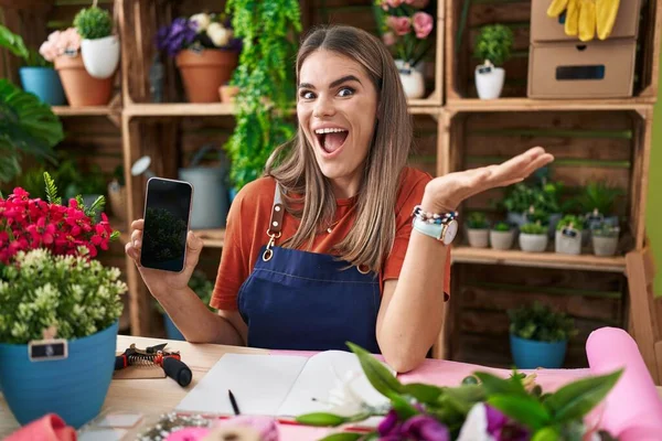 Hispanic young woman working at florist shop showing smartphone screen celebrating victory with happy smile and winner expression with raised hands