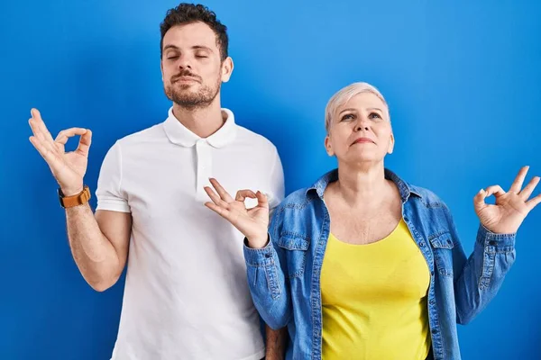 若いブラジル人の母親と息子が青い背景の上に立ってリラックスし 目を閉じて指で瞑想のジェスチャーを行う笑顔 ヨガのコンセプト — ストック写真