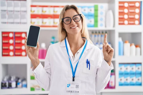 Young blonde woman working at pharmacy drugstore showing smartphone screen smiling with an idea or question pointing finger with happy face, number one