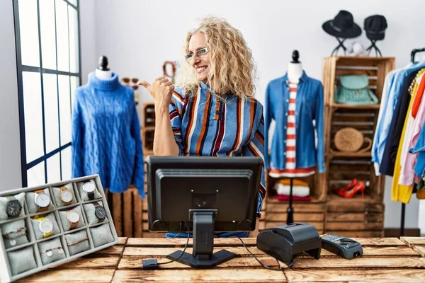 Middle age blonde woman working as manager at retail boutique smiling with happy face looking and pointing to the side with thumb up.